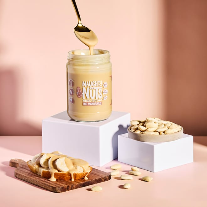 Bio Weißes Mandelmus wird mit einem Löffel aus einem Glas geschöpft, daneben Mandeln und ein Brot mit Bananen