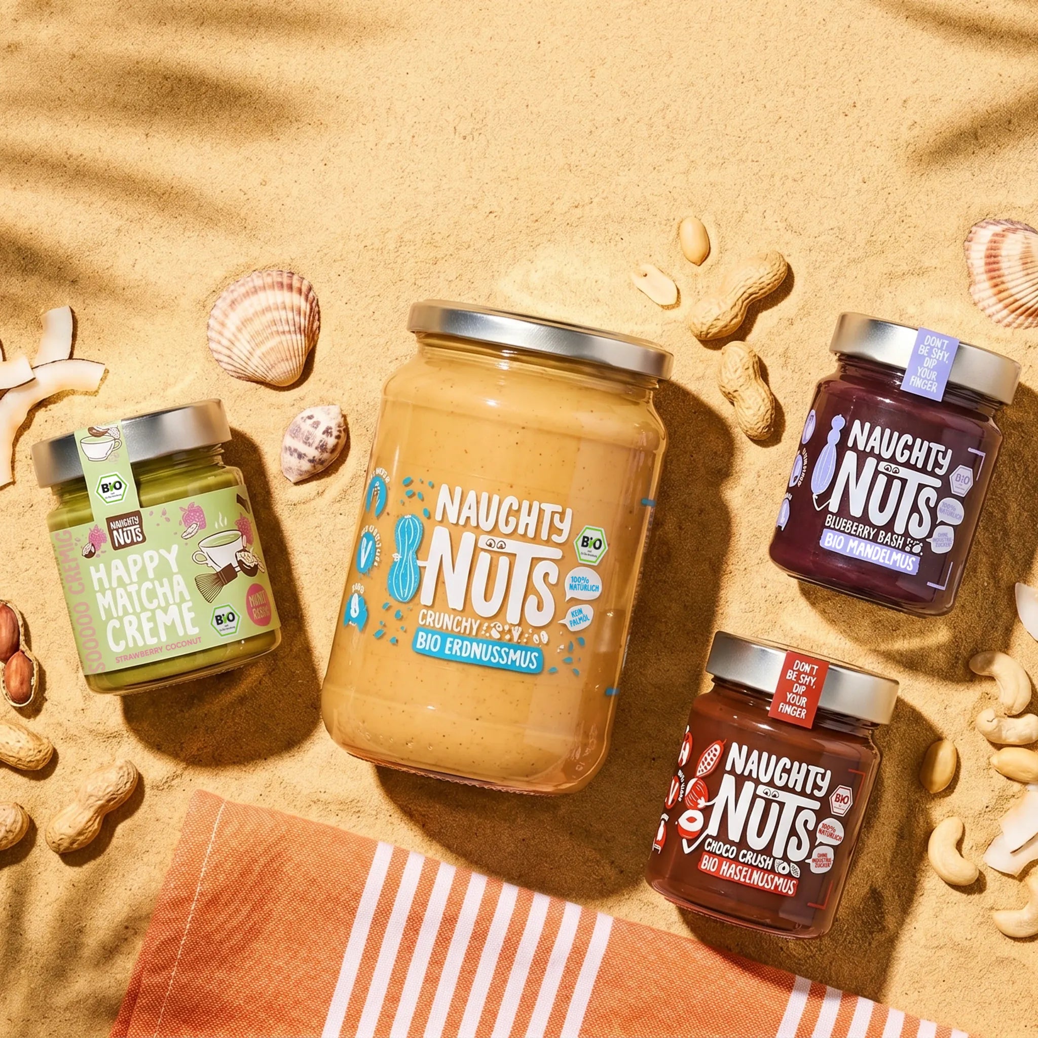 Vier Naughty Nuts Gläser auf Sand mit Muscheln, Erdnüssen, Cashews und Kokosnussstücken